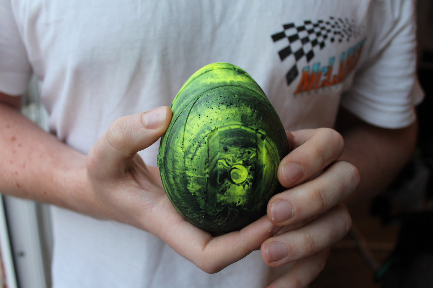 A person holding a small, vibrant neon yellow and black marbled Lando Norris inspired helmet in both hands. The helmet has a glossy finish and intricate marbling design.