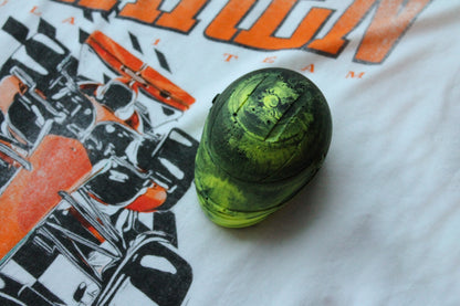 A top-down view of a vibrant neon yellow and black marbled mini helmet placed on a white t-shirt with an orange "McLaren" logo and an illustration of a race car. The helmet's unique marbling pattern and design details are highlighted against the shirt.