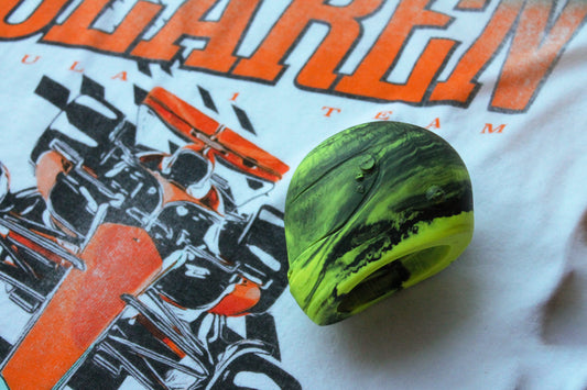 A vibrant neon yellow and black marbled mini helmet resting on a white t-shirt with a bold, orange "McLaren" logo and an illustration of a race car. The helmet's intricate design and marbling details are clearly visible.